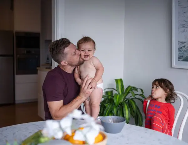 Een man die aan tafel zit en een kus geeft aan zijn baby. Naast hem zit zijn andere kind.